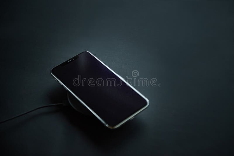 Mobile phone on wireless charger loading on black background with black screen, black wires in the corner of the photo to show contrast and simplicity of wireless charging nowadays. Mobile phone on wireless charger loading on black background with black screen, black wires in the corner of the photo to show contrast and simplicity of wireless charging nowadays.