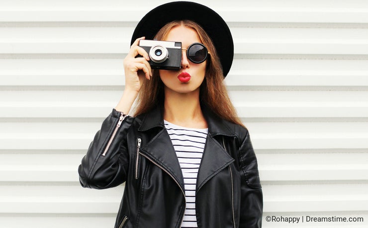 Fashion look, pretty cool young woman model with retro film camera wearing elegant black hat, leather rock jacket over white