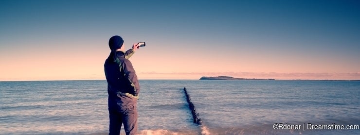 Tourist hold cellphone, take picture of autumn sea scenery at breakwater. Vivid and vignetting effect.