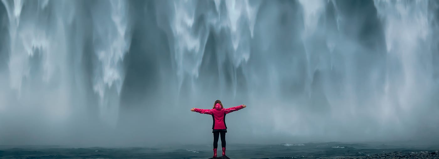 iceland landscape photo of brave girl who proudly standing with his arms raised in front of water wall of mighty waterfall famous