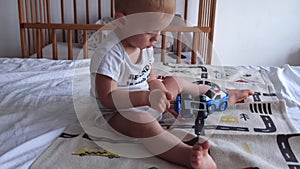 A 1 or 2 year old child plays with a screwdriver and toy police car in car repair sitting on bed at home