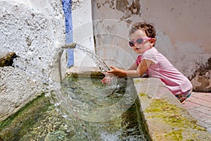 A 1-year-old girl refreshes herself with fresh water from a fountain in a small village on a summer day