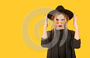 1 young white woman in a black jacket and hat, sunglasses  on a yellow background