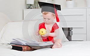 10 months old baby boy in graduation hat reading books and holding big apple