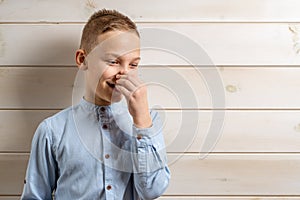 A 10-year-old boy in a blue shirt smiles on a light wooden background and covers his nose with his hands because of an unpleasant