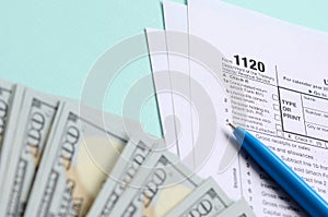 1120 tax form lies near hundred dollar bills and blue pen on a light blue background. US Corporation income tax return