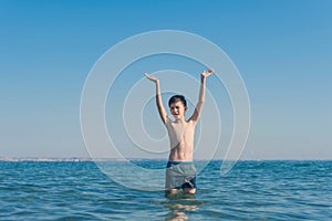 13 years old boy raises his hands standing up in the sea waves. Concept of family summer vacation