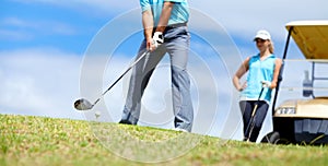 The 18th Hole - Make it count. A young female golfer leaning against a golf cart and watching her male partner play a