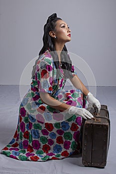 1950`s woman in a colourful dress crouching beside a suitcase against a white backdrop
