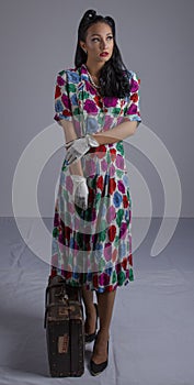 1950`s woman in a colourful dress crouching beside a suitcase against a white backdrop