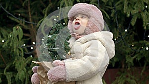 A 2-3 year old girl at the Christmas market holding a small Christmas tree. A child catches the falling snowflakes with