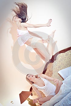2 beautiful happy women in pajamas one of them jumping, while the other looking at her on the bed