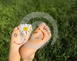 2 cute faces are drawn on the bare feet of a child lying on the grass