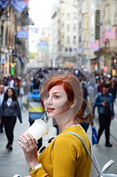2-cute girl with red hair drinks coffee from a paper cup with a straw on a crowded street and smiles.jpg