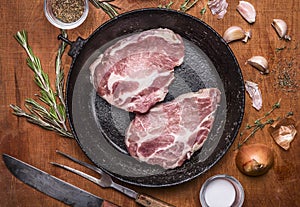 2 raw fresh pork steaks in old cast-iron frying pan with Onion garlic and herbs on rustic wooden background top view close up