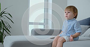A 2 years old boy sits on a sofa and watches TV while sitting with the remote control in his hands. View cartoons and TV