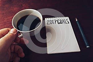 2017 goals list with notebook, man`s hand with cup of coffee on wooden table