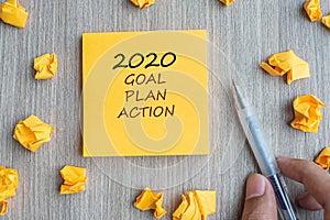 2020 Goal, Plan, Action word on yellow note with Businessman holding pen and crumbled paper on wooden table background. New Year