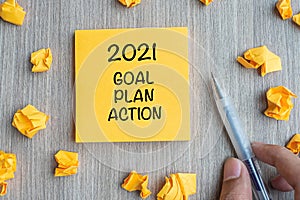 2021 Goal, Plan, Action word on yellow note with Businessman holding pen and crumbled paper on wooden table background. New Year