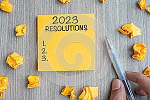 2023 RESOLUTIONS word on yellow note with Businessman holding pen and crumbled paper on wooden table background. New Year start,