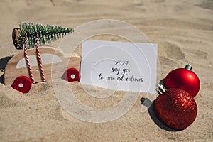 2024 SAY YES TO NEW ADVENTURES text on paper greeting card on background sandy beach sun coast. Christmas balls Santa