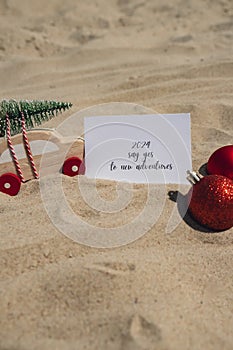 2024 SAY YES TO NEW ADVENTURES text on paper greeting card on background sandy beach sun coast. Christmas balls Santa