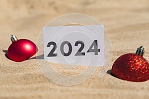2024 text on paper greeting card on background sandy beach sun coast. Christmas balls New Year New Me decoration. Summer