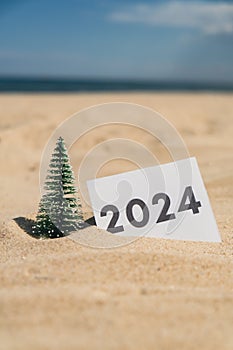 2024 text on paper greeting card on background sandy beach sun coast. Christmas tree New Year New Me decoration. Summer