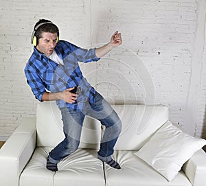 20s or 30s man jumped on couch listening to music on mobile phone with headphones playing air guitar