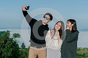 3 Asian friends group wearing sweaters, take a selfie phone, take pictures in the tourist area behind the fog and mountains with a