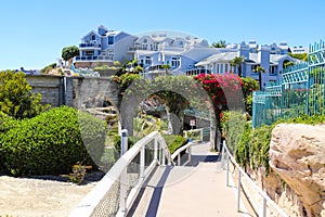 3 stone arches with lush green plants and red flowers down a staircase with a white hand rail with gray homes along the mountain