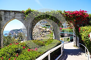 3 stone arches with lush green plants and red flowers down a staircase with a white hand rail with gray homes along the mountain