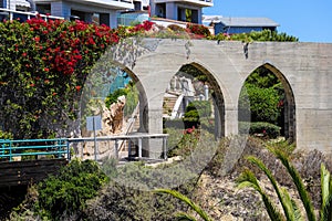 3 stone arches with lush green plants and red flowers down a staircase with a white hand rail with gray homes along the mountain