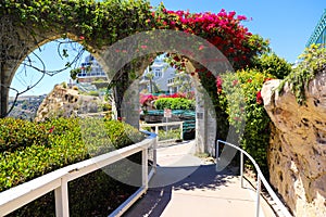 3 stone arches with lush green plants and red flowers down a staircase with a white hand rail with gray homes along the mountain