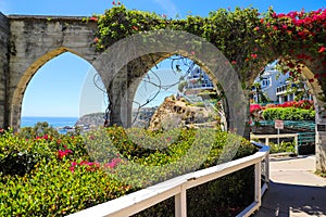 3 stone arches with lush green plants and red flowers down a staircase with a white hand rail with gray homes along the mountain