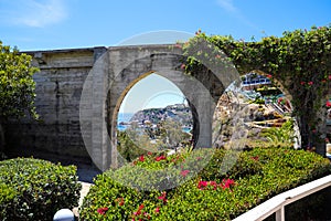 3 stone arches with lush green plants and red flowers down a staircase with a white hand rail with gray homes along the mountain