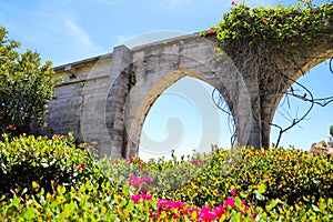 3 stone arches with lush green plants and red flowers down a staircase with a white hand rail with gray homes along the mountain