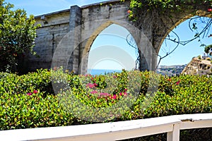 3 stone arches with lush green plants and red flowers down a staircase with a white hand rail with gray homes along the mountain