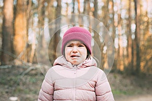 A 3-year-old girl in a pink jacket and a pink hat stands in the forest and cries