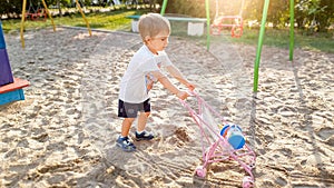 3 years old little boy with toy pram for dolls playing on the playground at park