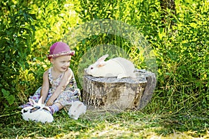 3 years old small girl in gray dress and pink hat sits on ground with four rabbits around. One big white easter rabbit sits on
