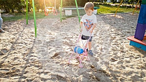 3 years old toddler boy playing with pram for dolls on the playground