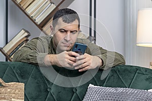 A 30-35-year-old man of oriental appearance looks through messages and videos on a smartphone while sitting behind a sofa.