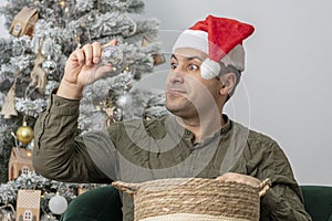 A 30-35-year-old man in a red hat looks at Christmas toys and balls from a wooden basket.
