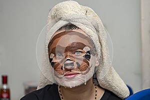A 30-35 year old woman sits at a table with a towel wrapped around her hair, with dog face makeup on her face.