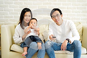 30s happy Asian family are watching TV and smiling while sitting on couch at home. Father is holding remote control in