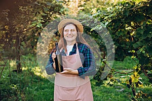30s Woman in hat tasting red wine in vineyard. Portrait of pretty young woman holding bottle of wine on sunny day