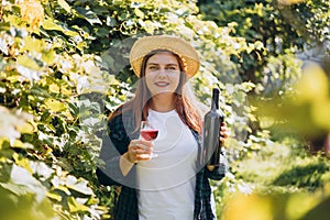 30s Woman in hat tasting red wine in vineyard. Portrait of pretty young woman holding glass of wine and bottle of wine