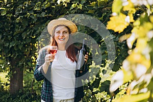 30s Woman in hat tasting red wine in vineyard. Portrait of pretty young woman holding glass of wine and bottle of wine