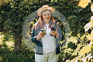 30s Woman in hat tasting red wine in vineyard. Portrait of pretty young woman holding smartphone and bottle of wine on